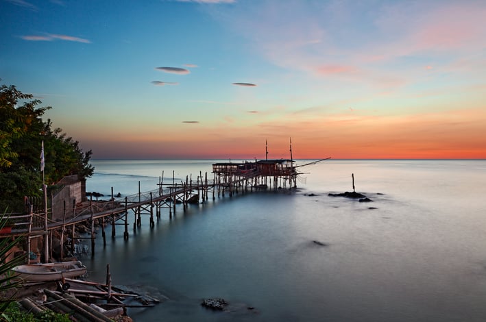 A trabocco at sunset in Chieti province, Abruzzo, with its long wooden walkway extending over still water.
