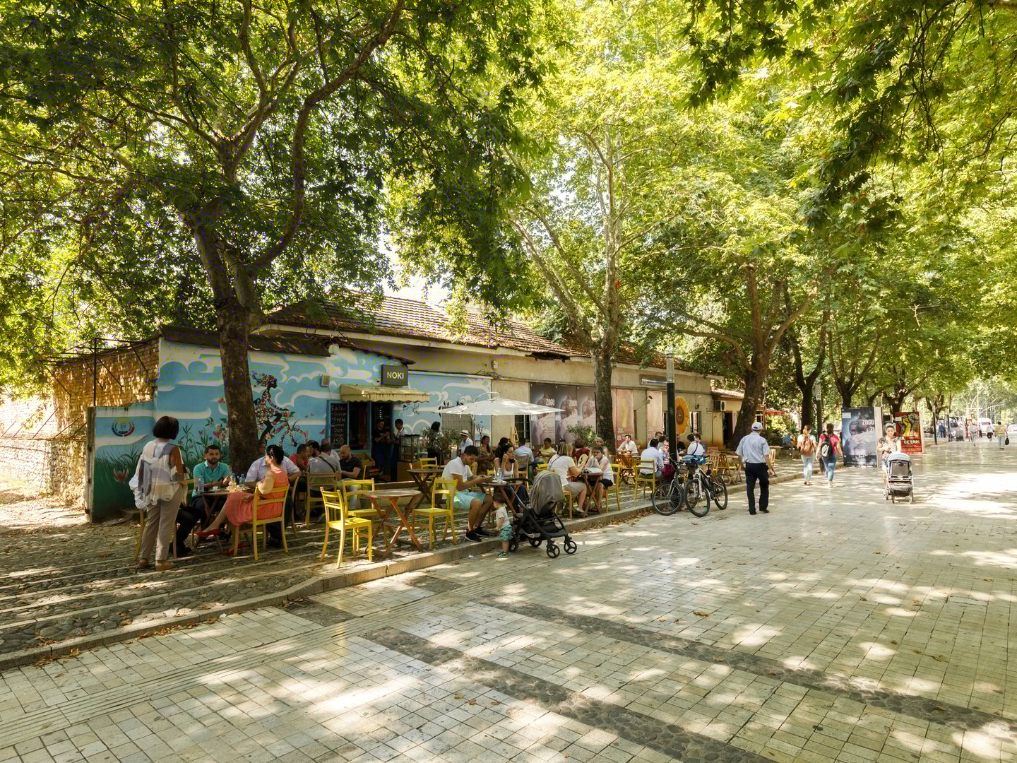 People walking along a wide pedestrian street with a cafe under the trees