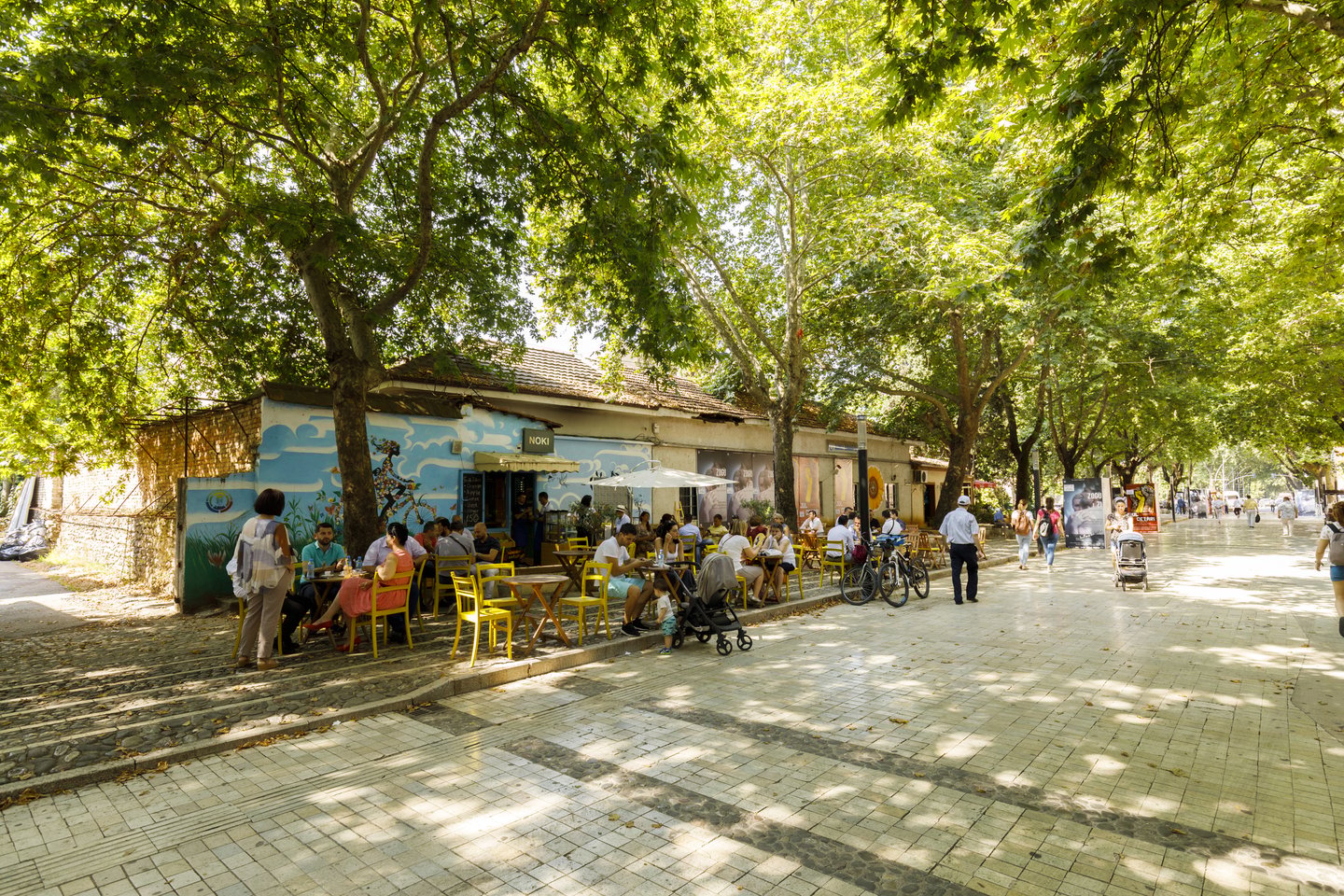 People walking along a wide pedestrian street with a cafe under the trees