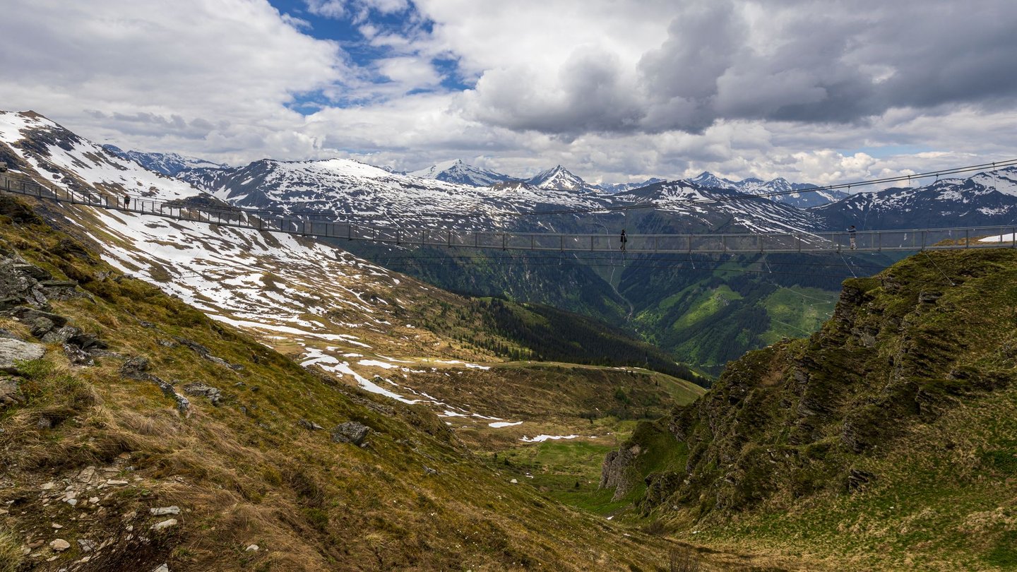 People walking on the Stubnerkogel Suspension Bridge with snow-capped mountains in the background.