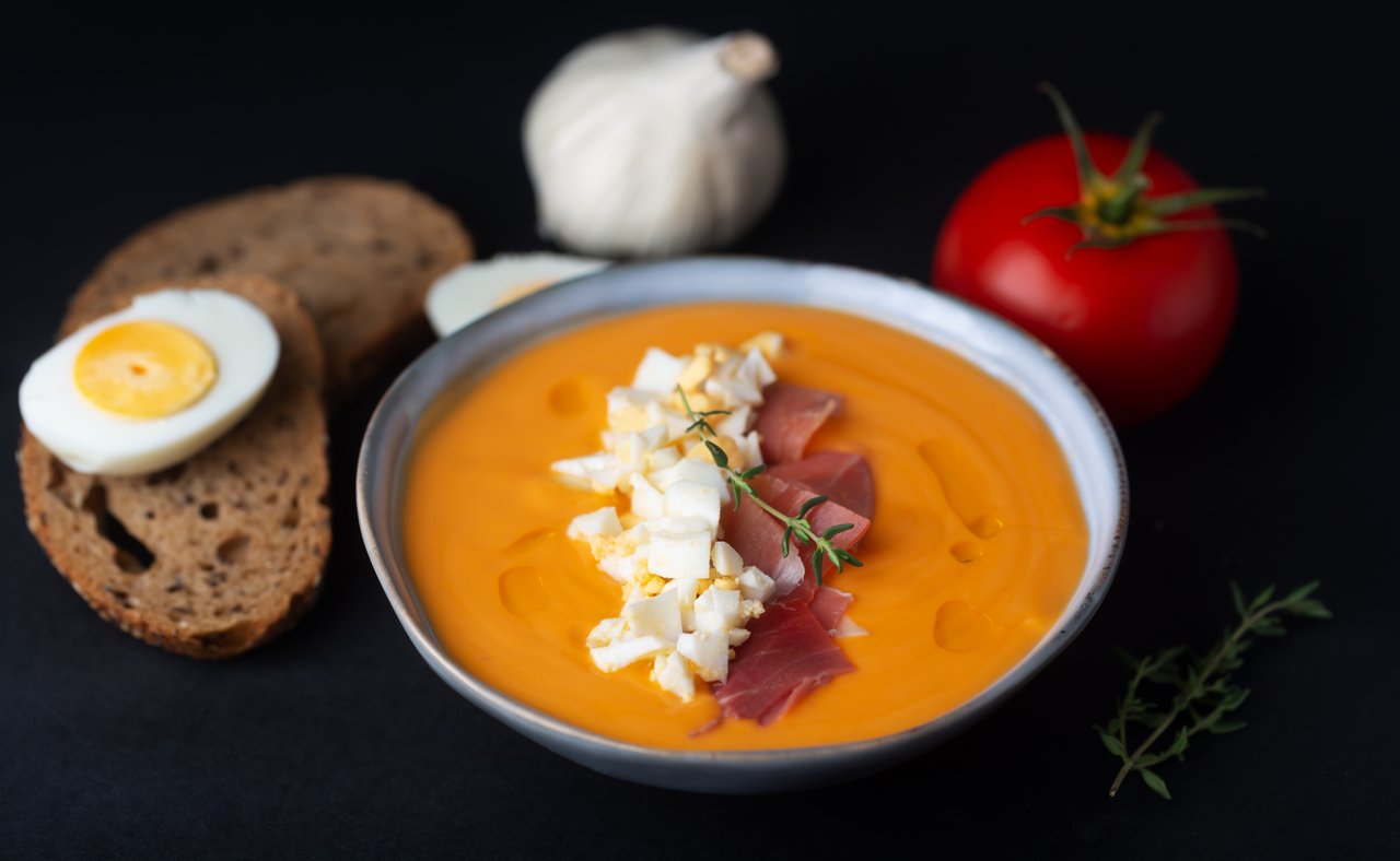 A bowl of salmorejo soup with the ingredients in the background