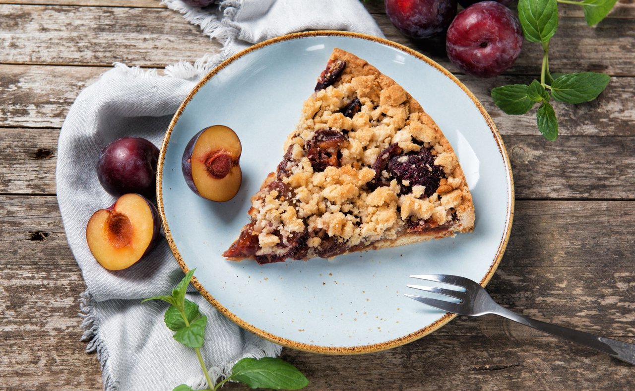A piece of Zwetchgenkuchen on a plate with plum in the background