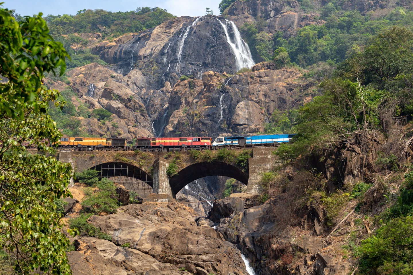 A colourful train on an old bridge bridge with the Dudhsagar Falls in the background.