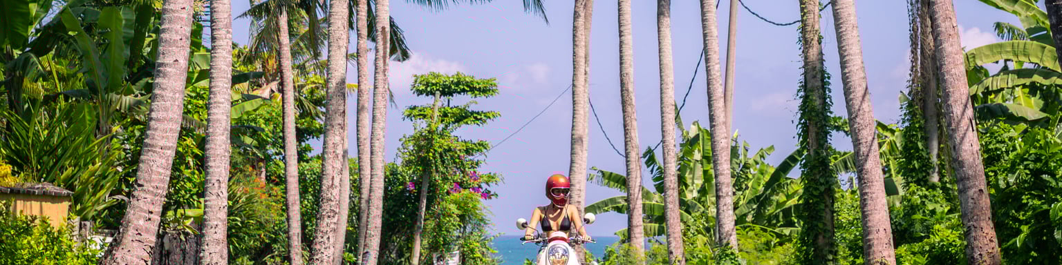 A young woman riding a scooter between palm trees in Bali.
