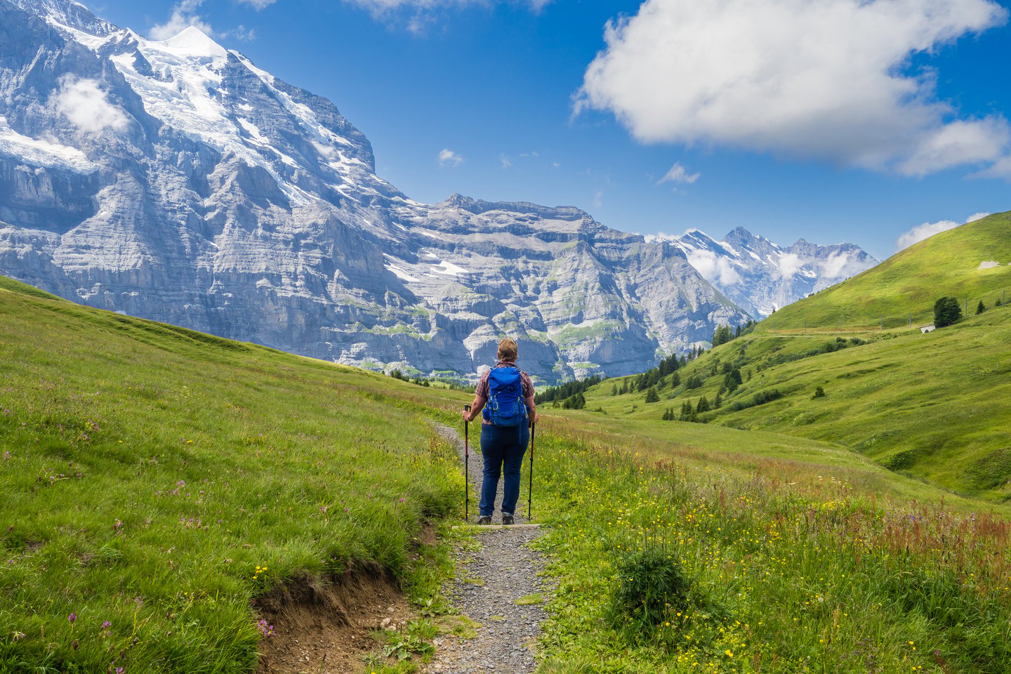 A woman hiking on the Eiger Trail carrying a backpack and poles with the mountains in front of her