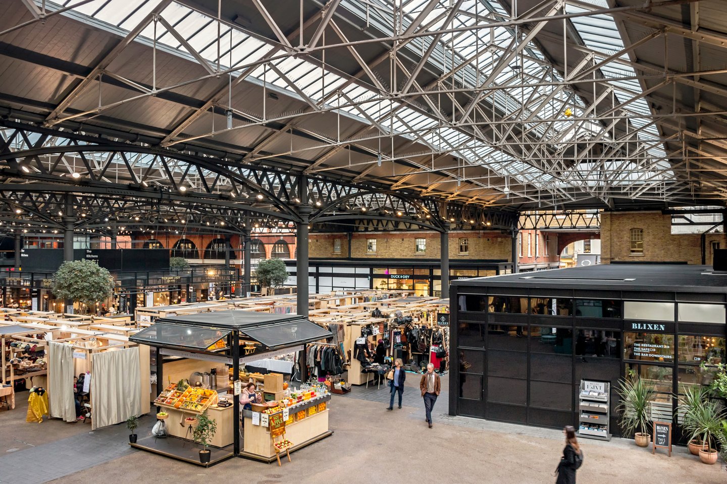People walking between stalls selling clothes and juice inside Spitalfields Market, London