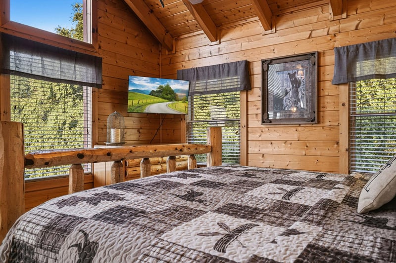 Wake up surrounded by warm wood and sunlight streaming through multiple windows framing leafy green views. Your rustic log bed and cozy quilted linens make mornings here feel like a peaceful mountain 