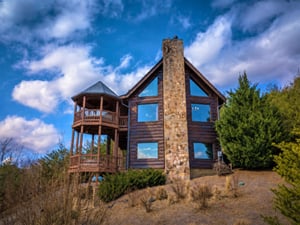 A striking mountain lodge with timber siding and stone chimney, featuring dramatic vaulted windows and multilevel decks, nestled among evergreens beneath expansive blue skies.