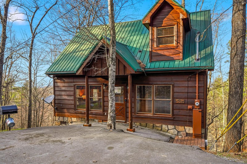 Charming log cabin retreat nestled among towering trees, featuring rustic wood siding, green metal roofing, and welcoming covered entrance with convenient parking.