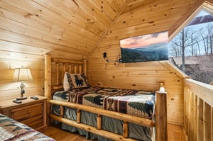Your cozy loft bedroom features rustic log walls, quilted bedding, and a large window showcasing breathtaking mountain views.