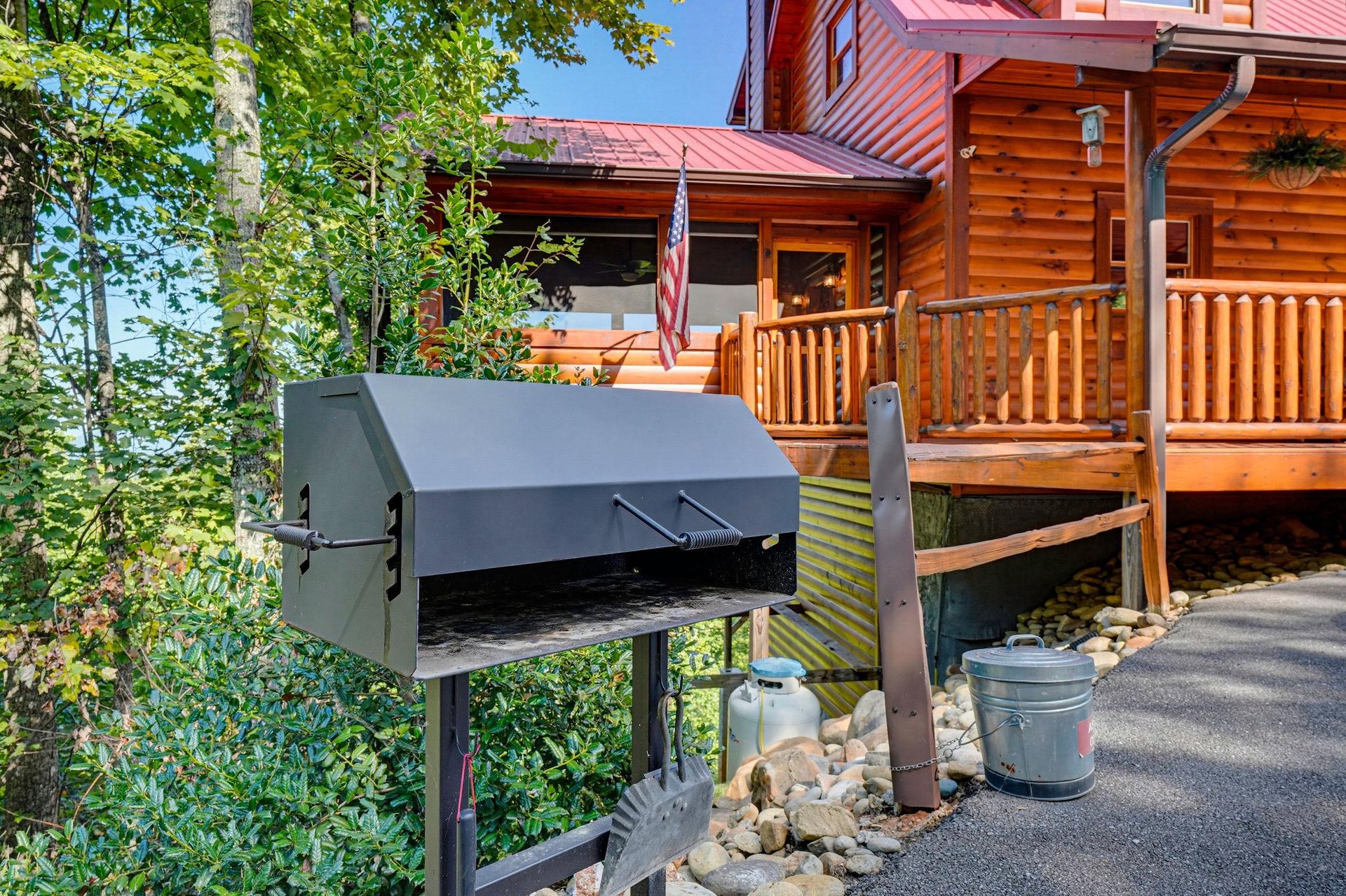 Charcoal grill stationed at the entrance of a rustic log cabin set among birch trees and lush greenery on a sunny day.