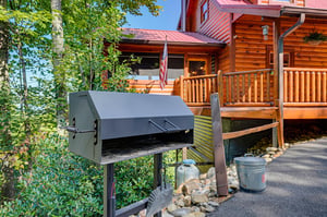 Charcoal grill stationed at the entrance of a rustic log cabin set among birch trees and lush greenery on a sunny day.