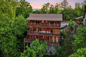 Multilevel mountain cabin surrounded by lush forest canopy at twilight, featuring wraparound decks on each floor.