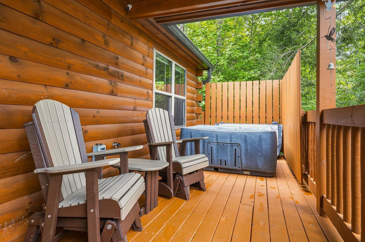 Your private covered deck awaits with Adirondack chairs, a footstool for lounging, and your own hot tub overlooking lush summer greenery—perfect for unwinding after a day of exploring.