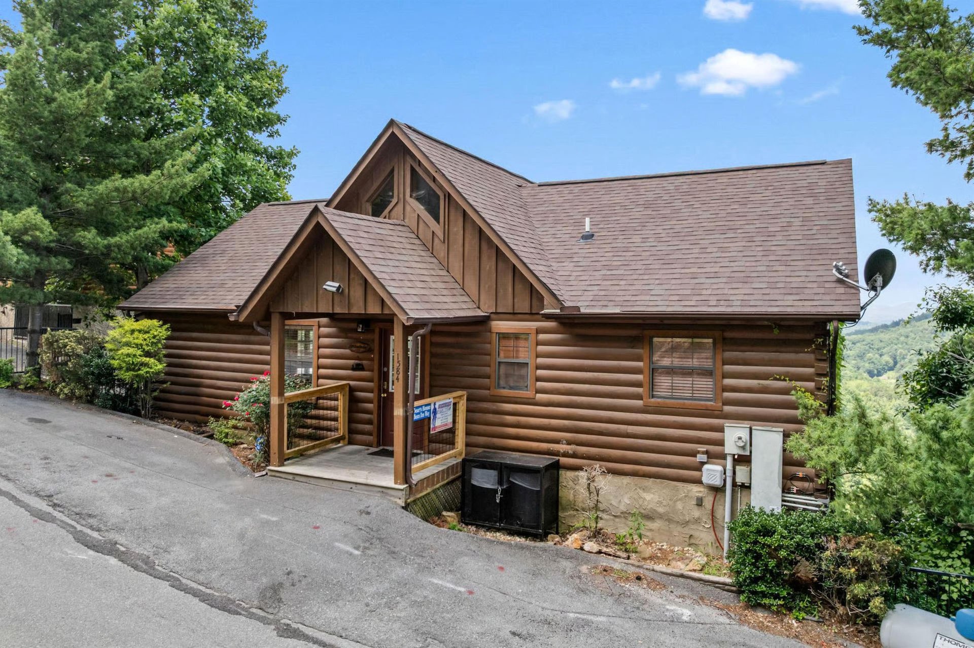A charming log cabin rental nestled in the mountains, featuring rustic wood architecture and surrounded by lush trees with scenic valley views in the background.