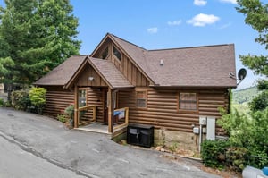 A charming log cabin rental nestled in the mountains, featuring rustic wood architecture and surrounded by lush trees with scenic valley views in the background.