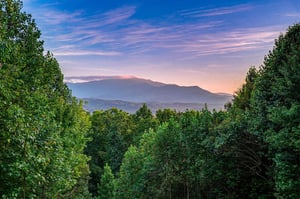 Stunning mountain vistas frame the property, with layered peaks rising beyond lush forest canopy. A tranquil natural setting where vibrant sunsets paint the sky in soft pinks and blues.