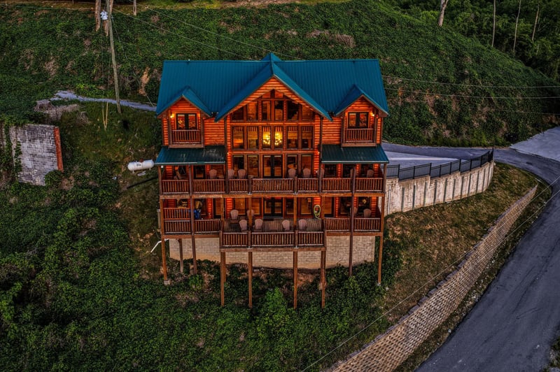 A striking mountain cabin perched on a hillside, featuring expansive log construction with a distinctive blue metal roof and multiple private balconies overlooking the surrounding forested landscape.