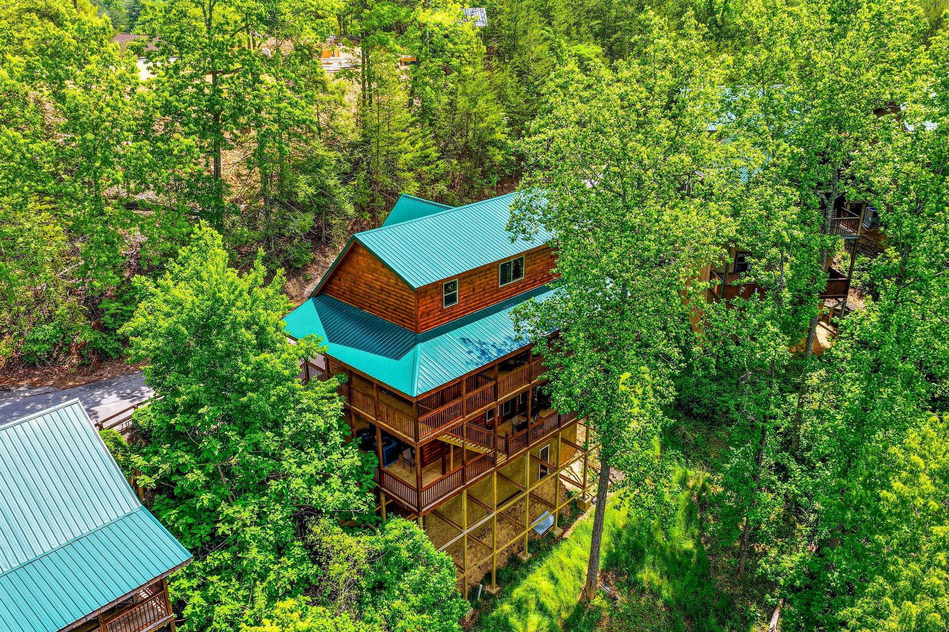 Aerial view of a charming log cabin nestled in lush forest canopy with distinctive turquoise metal roofing and multiple outdoor decks.