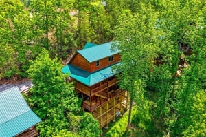 Aerial view of a charming log cabin nestled in lush forest canopy with distinctive turquoise metal roofing and multiple outdoor decks.