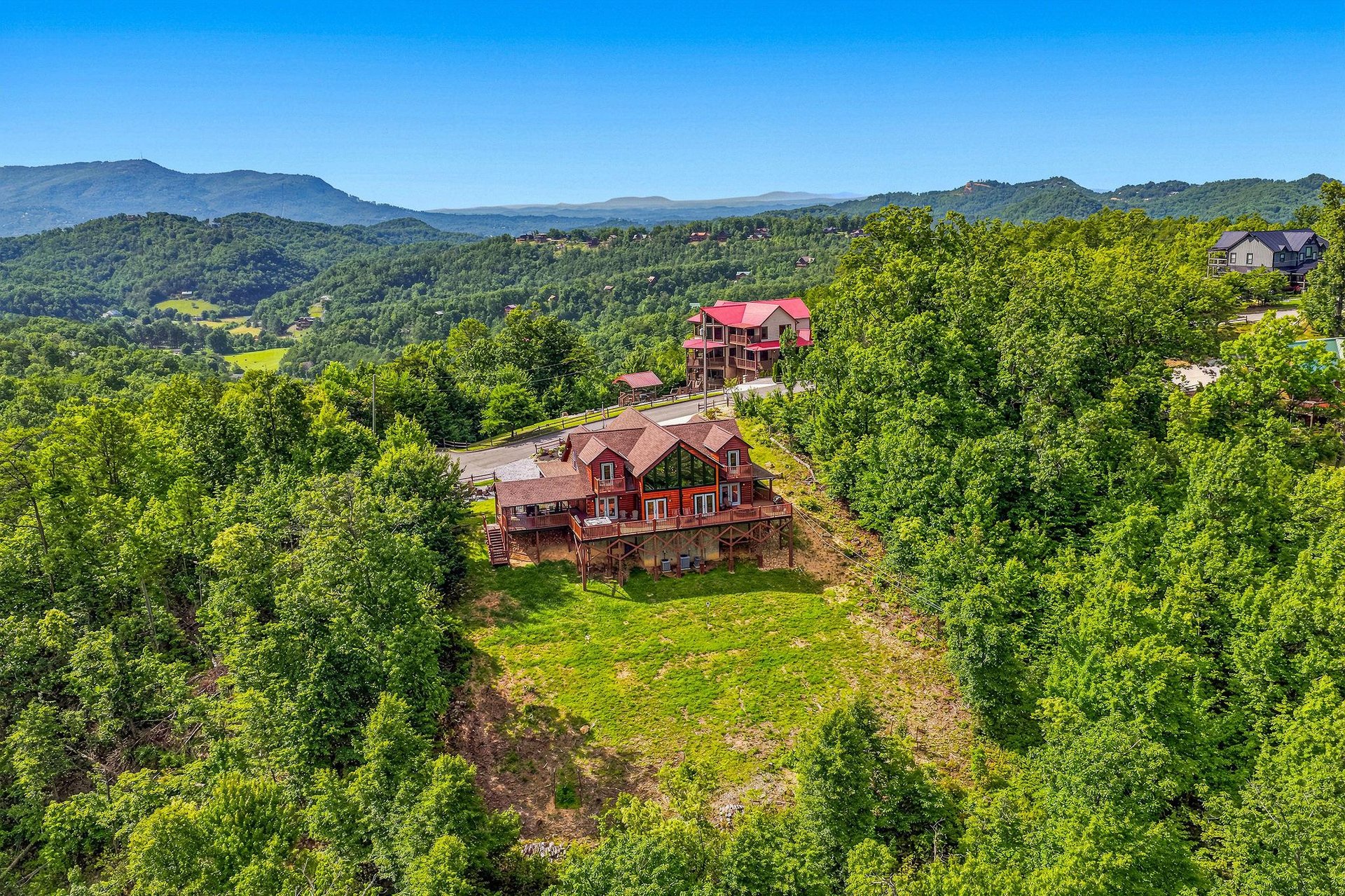 A charming red barnstyle vacation home sits on a hillside surrounded by lush forests and sweeping mountain vistas under a bright blue sky.