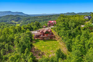 A charming red barnstyle vacation home sits on a hillside surrounded by lush forests and sweeping mountain vistas under a bright blue sky.