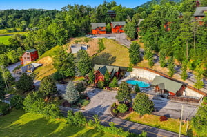 An aerial view showcases the vacation rental property nestled in lush mountain scenery, featuring multiple cabinstyle buildings, a refreshing pool area, and ample parking spaces surrounded by natural