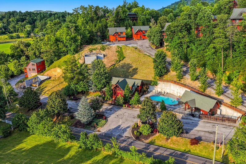 An aerial view showcases the vacation rental property nestled in lush mountain scenery, featuring multiple cabinstyle buildings, a refreshing pool area, and ample parking spaces surrounded by natural
