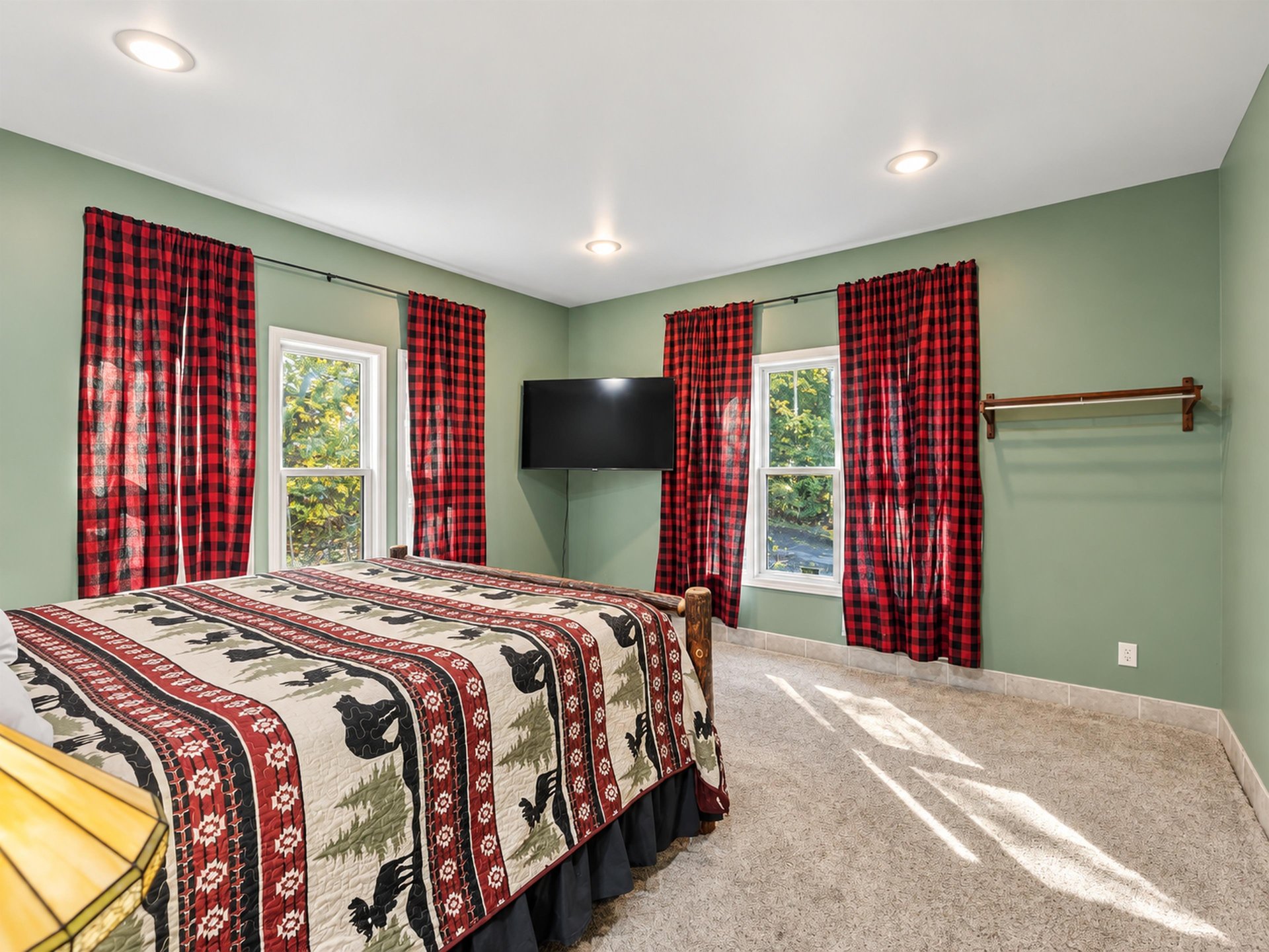 Unwind in this cozy bedroom with sagegreen walls and rustic touches. Sunlight streams through large windows, while charming plaid curtains frame peaceful wooded views.