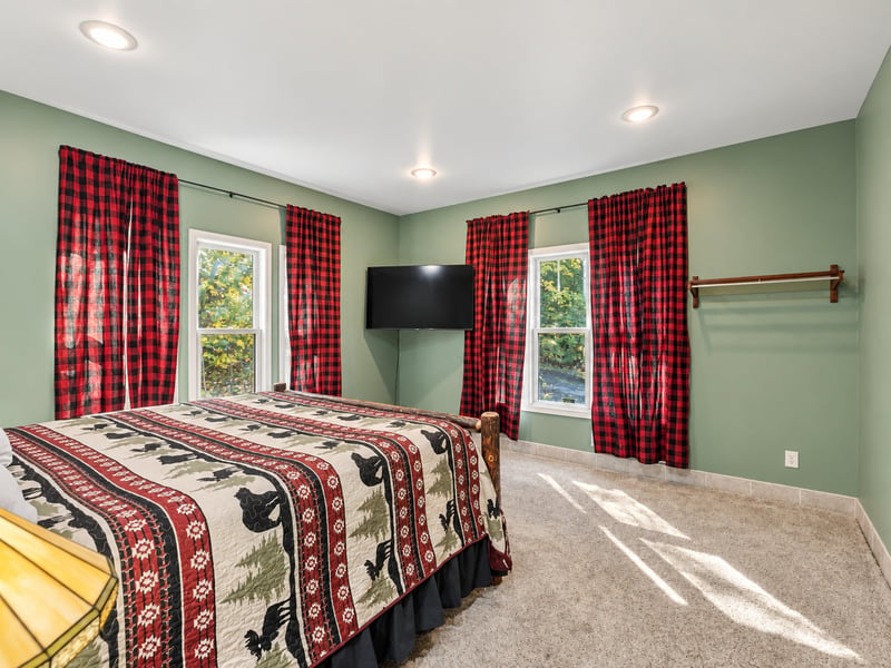 Unwind in this cozy bedroom with sagegreen walls and rustic touches. Sunlight streams through large windows, while charming plaid curtains frame peaceful wooded views.