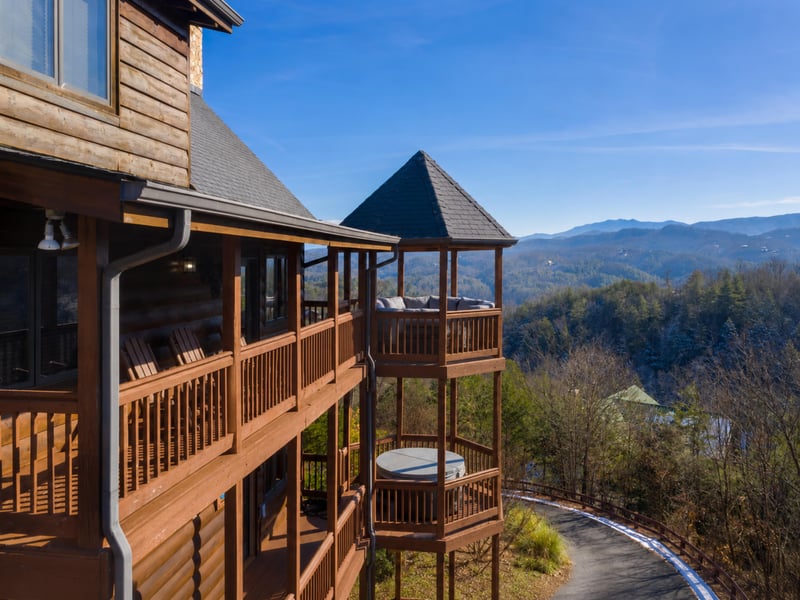 Mountain lodge exterior with multilevel wooden deck featuring cozy covered gazebo areas, a hot tub, and sweeping mountain vistas across forested terrain under clear blue skies.