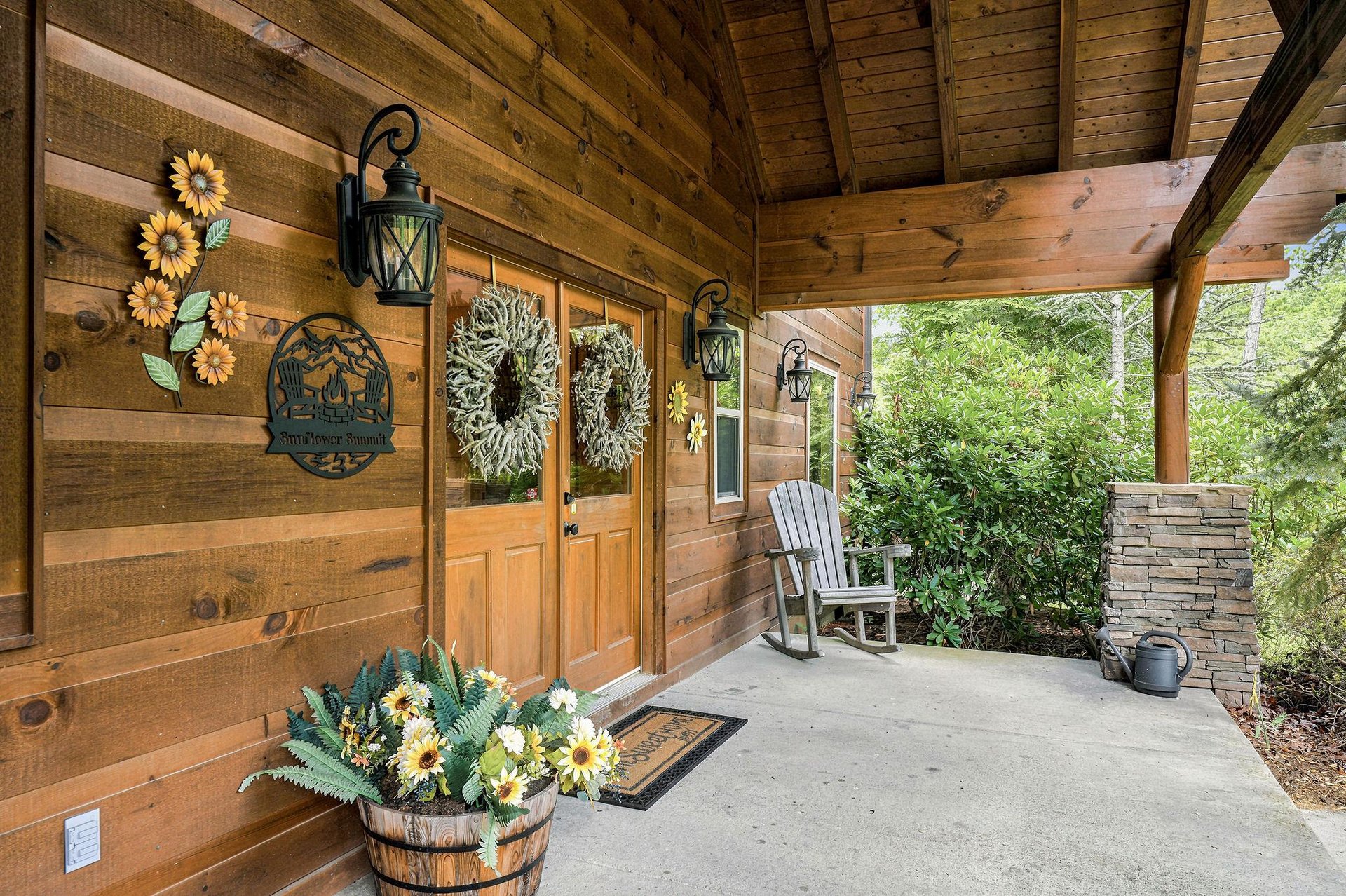 A warm woodpaneled porch welcomes guests with cheerful sunflower décor and a cozy rocking chair, surrounded by lush greenery and natural stone accents that frame the peaceful wooded setting.