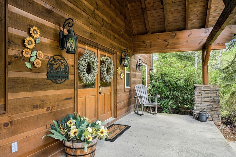 A warm woodpaneled porch welcomes guests with cheerful sunflower décor and a cozy rocking chair, surrounded by lush greenery and natural stone accents that frame the peaceful wooded setting.