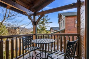 A covered balcony overlooks scenic mountain vistas and neighboring cabins, with a cozy bistro table inviting you to enjoy morning coffee or evening relaxation in the fresh air.
