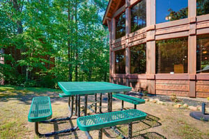 A teal picnic table sits ready in the wooded yard, inviting you to enjoy meals or morning coffee surrounded by tall, leafy trees and the peaceful quiet of nature.