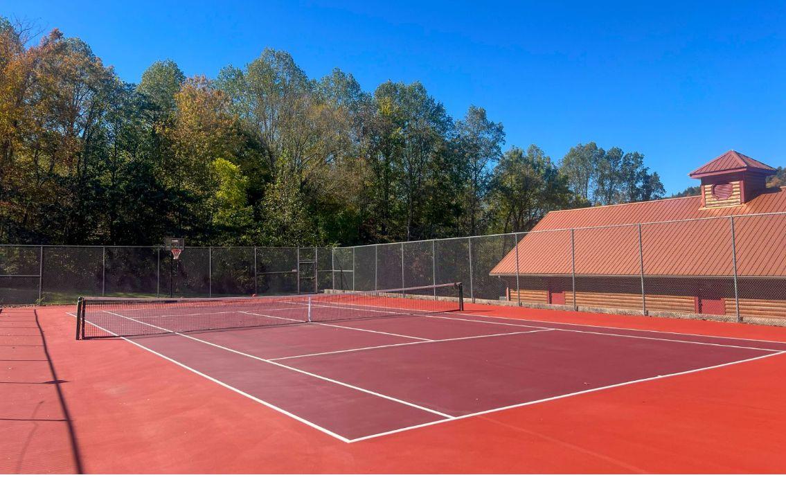 Professional tennis court with vibrant red surface surrounded by autumn foliage and rustic lodge architecture.