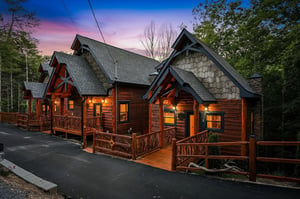 This charming log cabin awaits your arrival with warm interior lighting glowing through inviting windows, framed by rich wood siding, rustic railings, and towering trees at twilight.