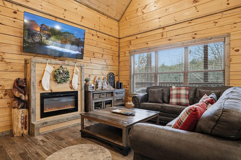 Unwind in your cozy living room with rustic wood walls, plush sectional seating, and a crackling fireplace creating the perfect mountain retreat atmosphere.