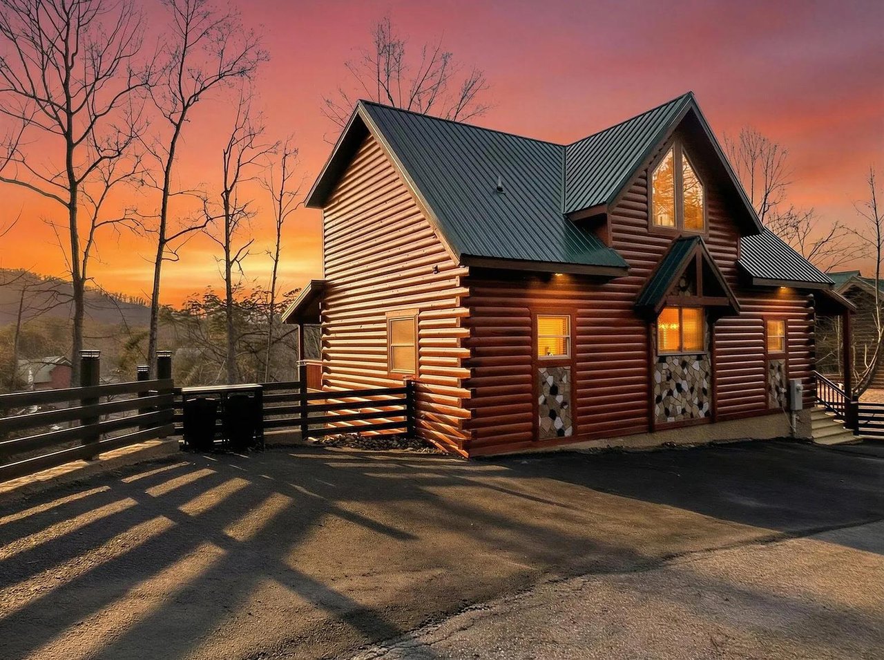 Modern log cabin exterior featuring warm lighting and dramatic sunset sky in scenic mountain setting.