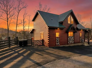 Modern log cabin exterior featuring warm lighting and dramatic sunset sky in scenic mountain setting.