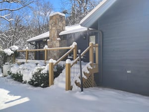 Snowcovered property entrance featuring stone chimney and wooden deck railing under winters pristine blanket.