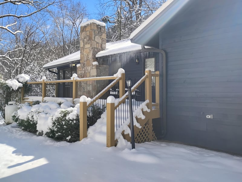 Snowcovered property entrance featuring stone chimney and wooden deck railing under winters pristine blanket.