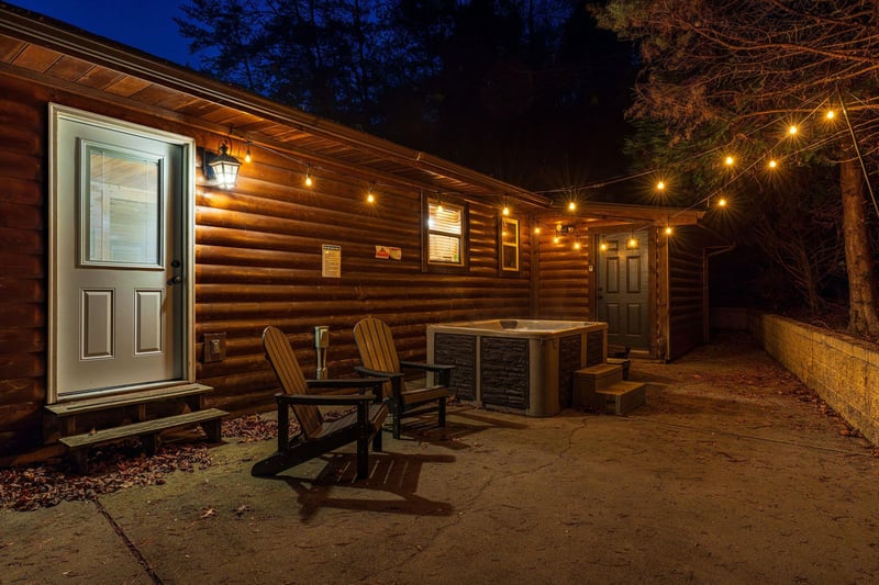 A cozy log cabin exterior with warm string lights creating an inviting nighttime ambiance. Relax in Adirondack chairs on the patio, or unwind in the hot tub under the stars.