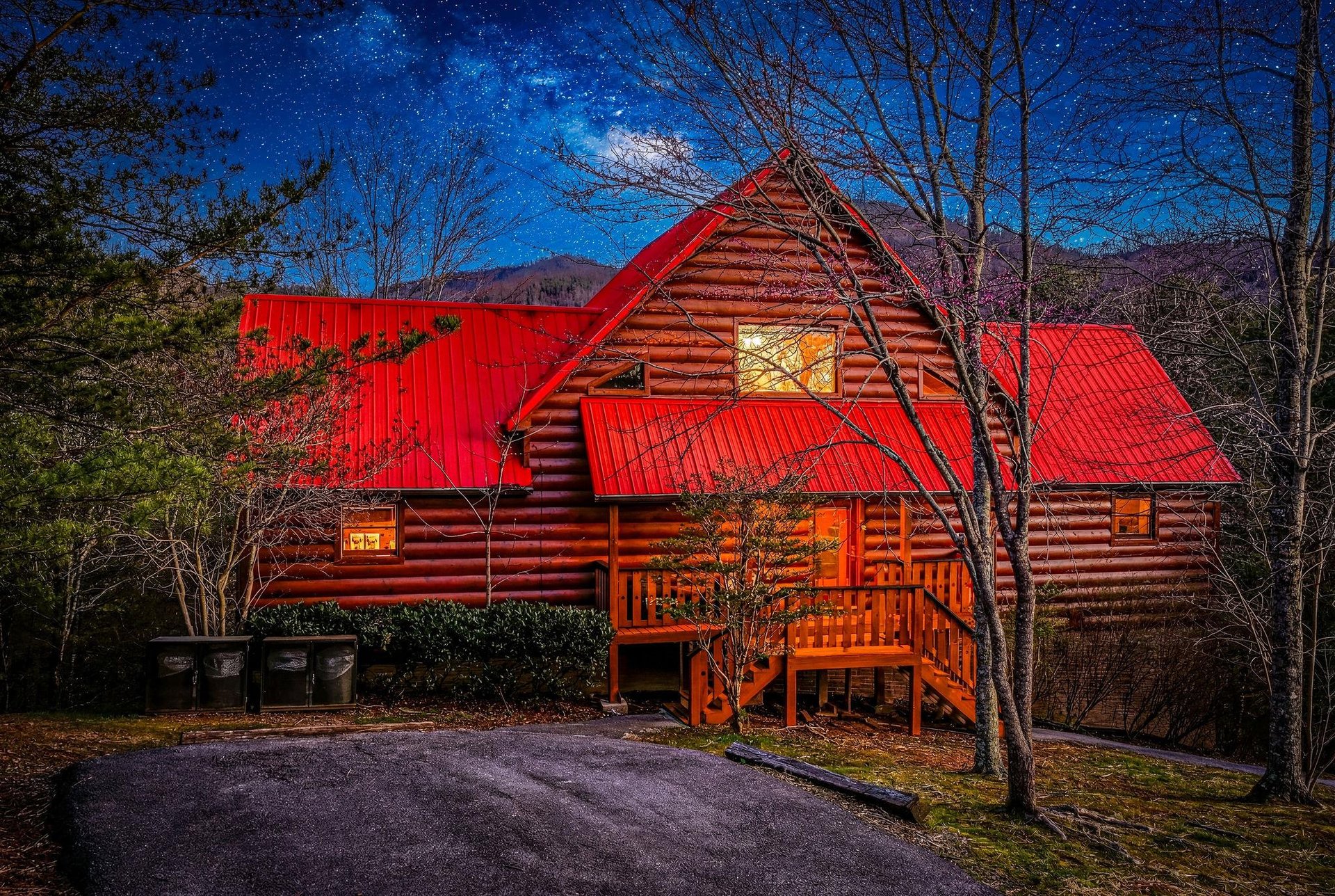 Rustic log cabin with red metal roof nestled among trees under starlit mountain sky, featuring warm glowing windows and welcoming front porch.