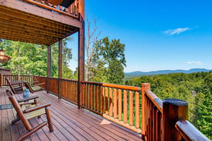 Breathtaking mountain vistas stretch endlessly from this inviting deck, where rustic wooden chairs await your morning coffee beneath sheltering cabin eaves surrounded by lush forest canopy.