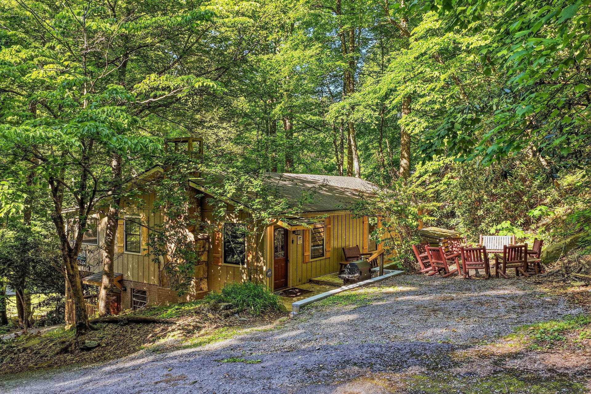 A welcoming cabin nestled beneath towering trees, with a gravel driveway and red porch seating ready for your arrival. This serene wooded retreat offers a peaceful escape immersed in nature.