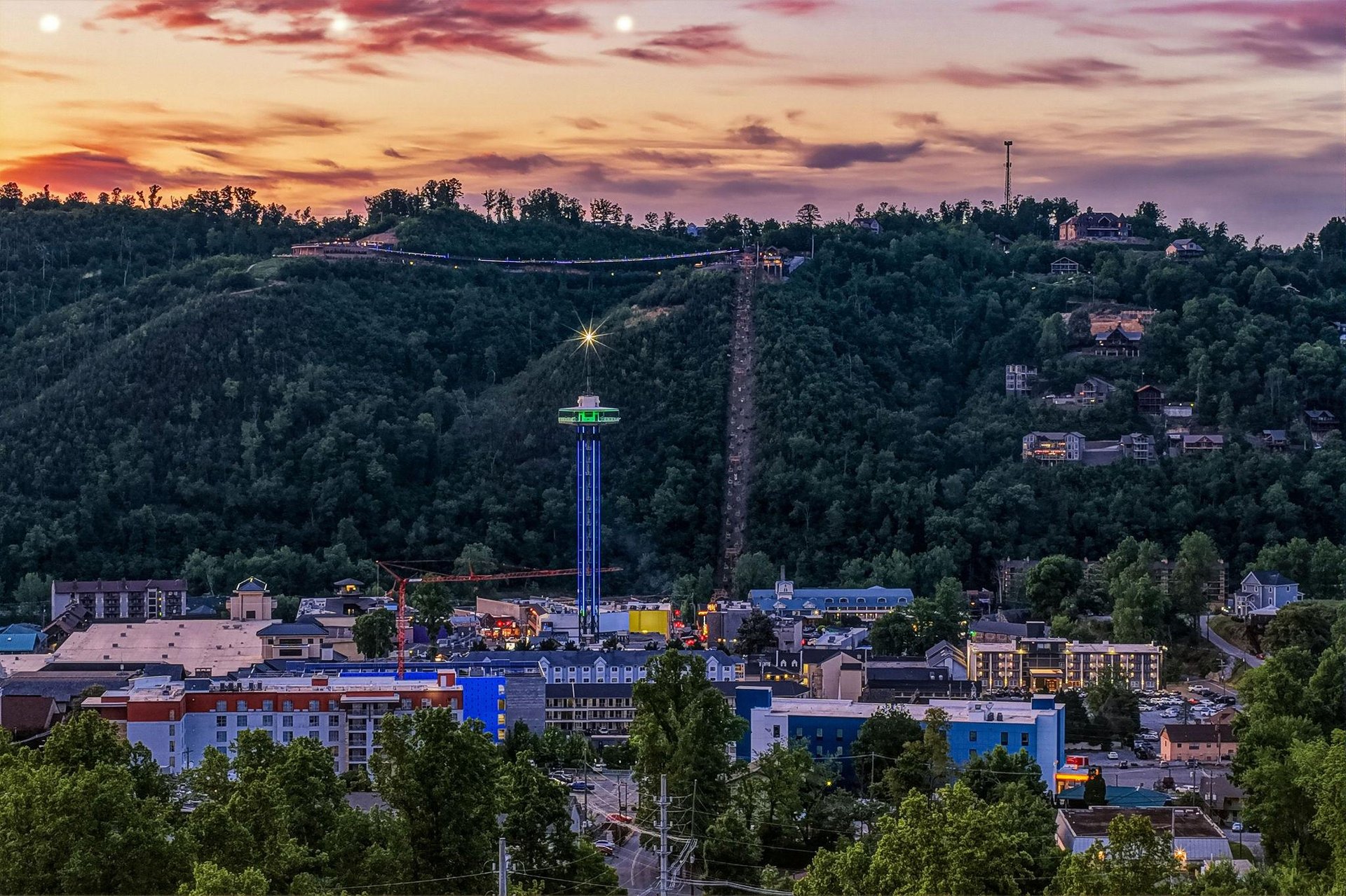 Hillside town at dusk with illuminated tower and residential buildings nestled among lush forested slopes beneath a dramatic sunset sky.