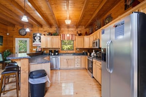 Your fully equipped cabin kitchen features warm wood cabinetry, granite counters, stainless appliances, and an island with seating—perfect for cozy homecooked meals under soaring beam ceilings.