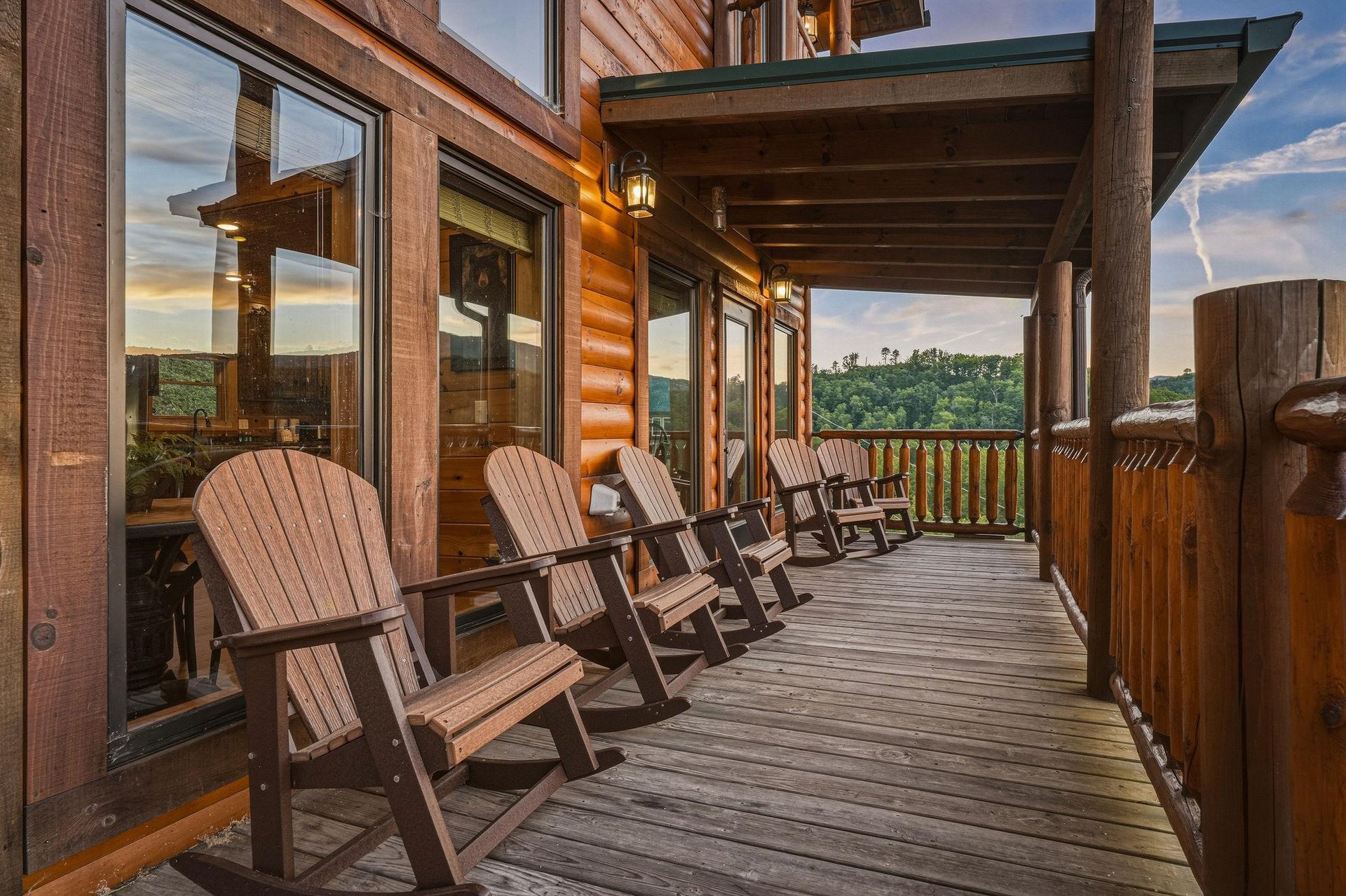 A row of rustic Adirondack chairs lines a spacious covered deck overlooking forested hillsides at twilight. Warm lanterns invite you to relax and soak in the mountain views.