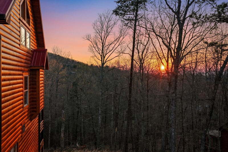 A rustic log cabin nestled among trees with sunset painting the sky in warm hues over distant mountains.